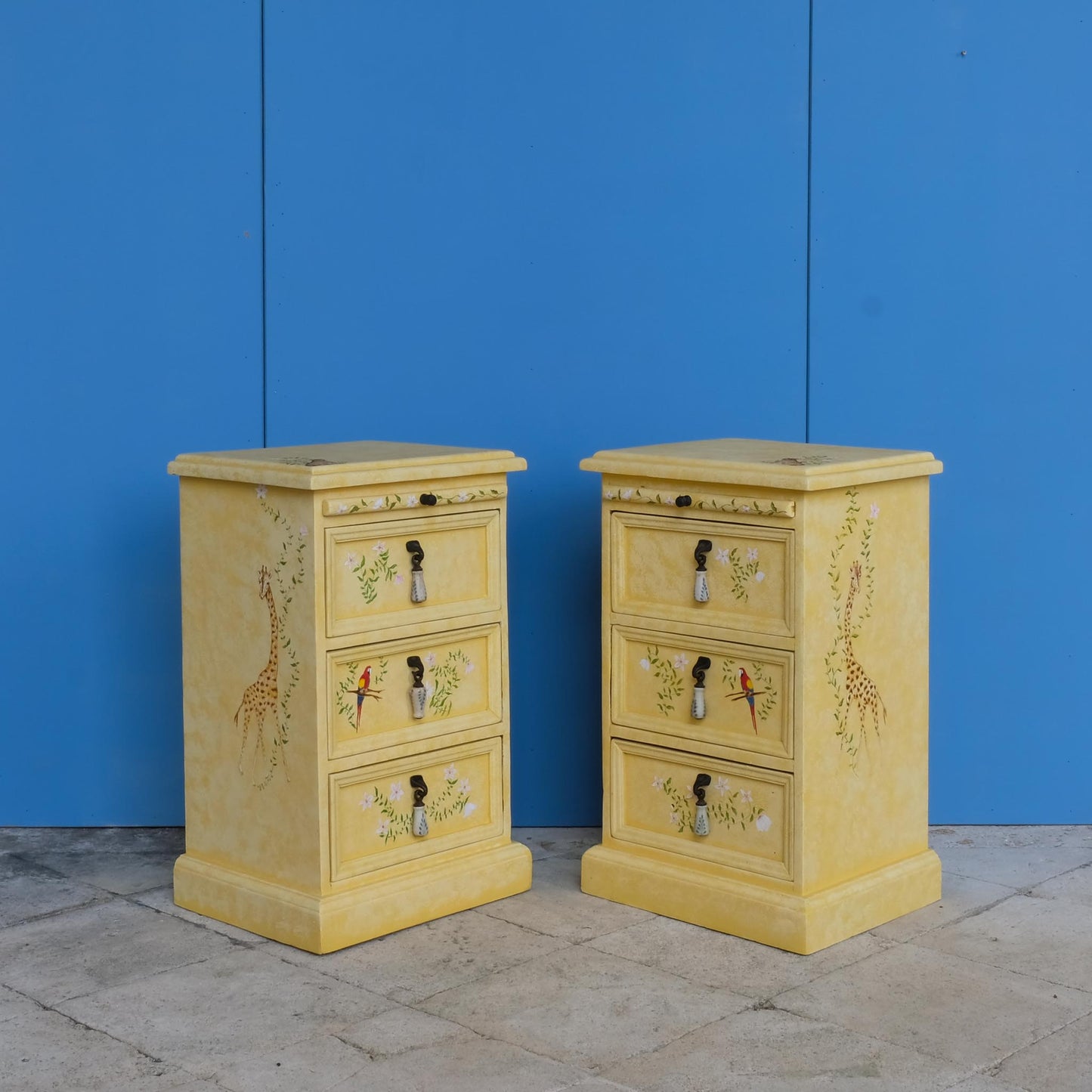 A stunning, unique matching pair of hand-painted chests of drawers, with brushing slides over three drawers. Finished in a fetching patinated yellow and decorated with exotic animals and Flora, in very good condition. Ideal for a children's bedroom or to liven up a sitting room. A unique pair in collaboration with Bunny Howe.