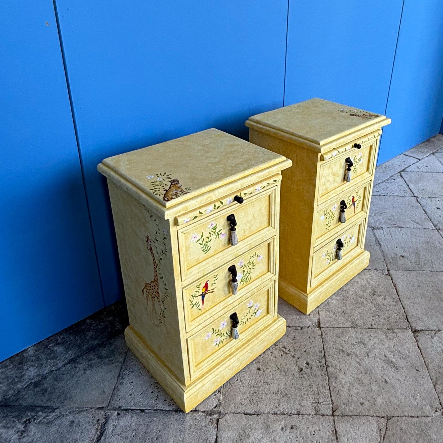 A stunning, unique matching pair of hand-painted chests of drawers, with brushing slides over three drawers. Finished in a fetching patinated yellow and decorated with exotic animals and Flora, in very good condition. Ideal for a children's bedroom or to liven up a sitting room. A unique pair in collaboration with Bunny Howe.